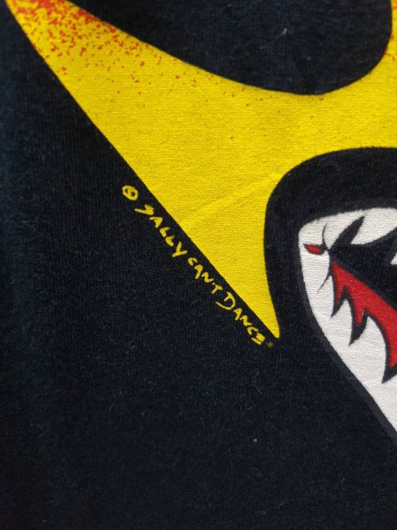 May include: Black t-shirt with a yellow and red shark tooth graphic. The text "Sally  Sant'Dng" is printed in yellow on the shirt.