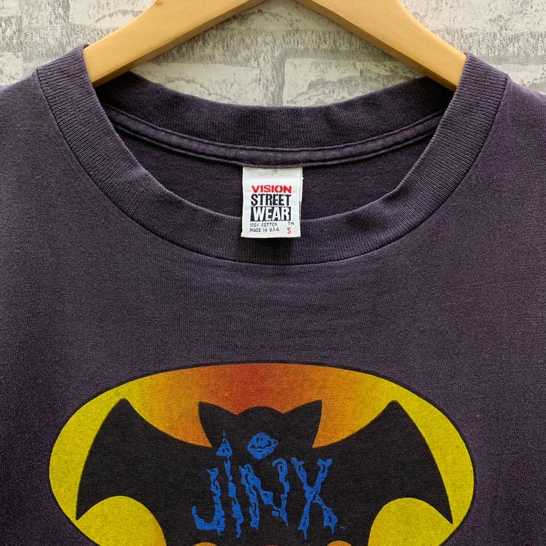 May include: A dark purple t-shirt with a yellow and orange bat graphic. The bat has a blue "JINX" written on it. The t-shirt has a "VISION STREET WEAR" tag on the front.