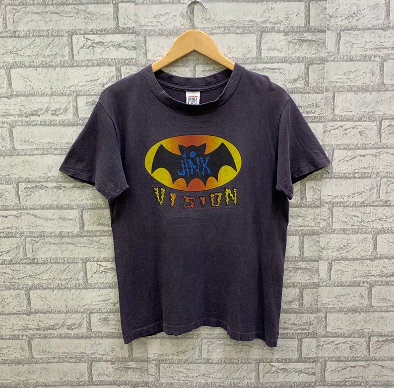 May include: A dark grey t-shirt with a yellow bat graphic and the text "JINX VISION" printed on the front.