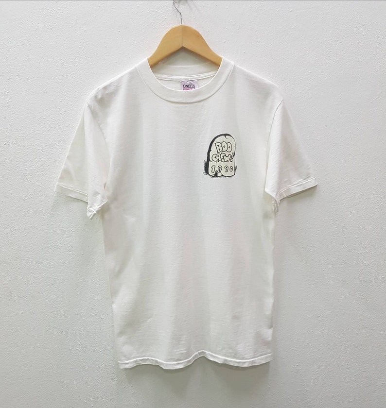 Puede incluir: Camiseta blanca de manga corta con un gr&aacute;fico en blanco y negro de una l&aacute;pida con el texto "B.D.D. Crew 1998" en ella.