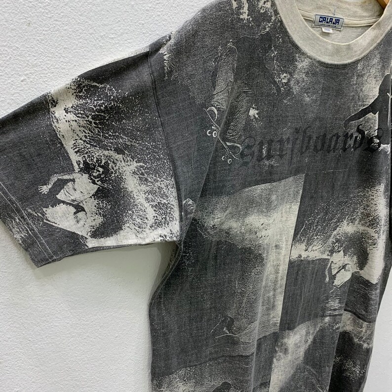 May include: Gray short-sleeved t-shirt with a white and gray all-over print of surfers and waves. The text "Surfboards" is printed in black on the front of the shirt.