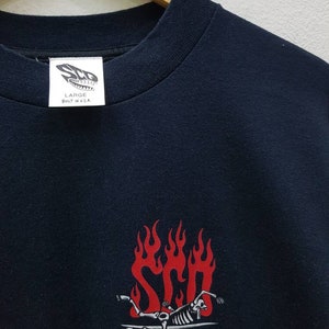 May include: Black t-shirt with a red and white graphic of a skeleton surfing on a surfboard with flames. The text "STP" is visible in the flames. The tag on the shirt reads "Large Built in U.S.A."