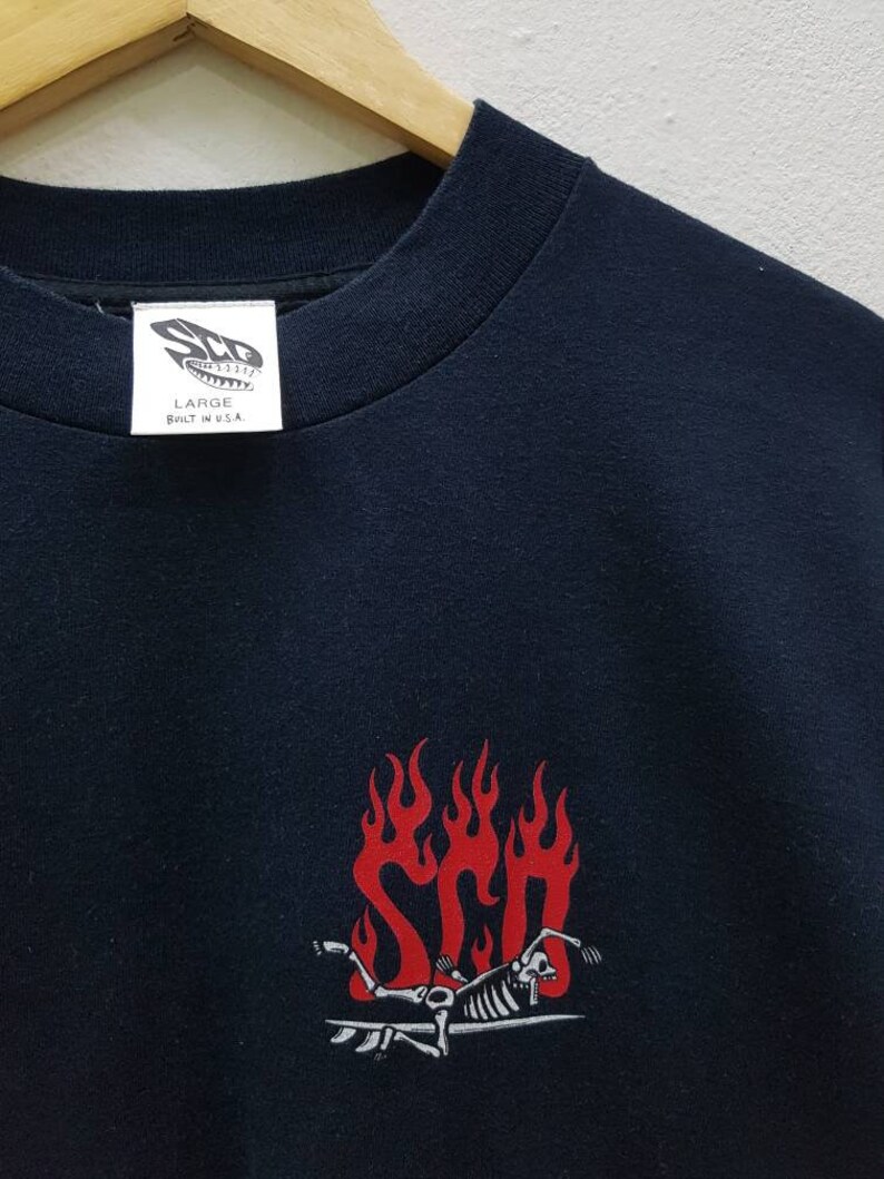 May include: Black t-shirt with a red and white graphic of a skeleton surfing on a surfboard with flames. The text "STP" is visible in the flames. The tag on the shirt reads "Large Built in U.S.A."