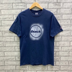 May include: A navy blue t-shirt with a white circle graphic featuring the word "FUCT" in all caps. The shirt has a Champion logo on the right sleeve.