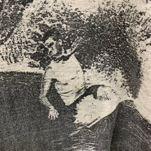 May include: A black and white graphic print of a person surfing on a wave. The print is on a gray t-shirt.