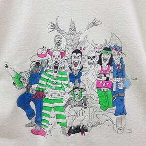 Puede incluir: Una camiseta blanca con una ilustraci&oacute;n en blanco y negro de un grupo de personajes espeluznantes. Los personajes incluyen un payaso, un vampiro, una bruja, un zombi y un monstruo. La ilustraci&oacute;n est&aacute; rodeada de un fondo blanco y negro con un &aacute;rbol y una l&aacute;pida que dice "RIP".