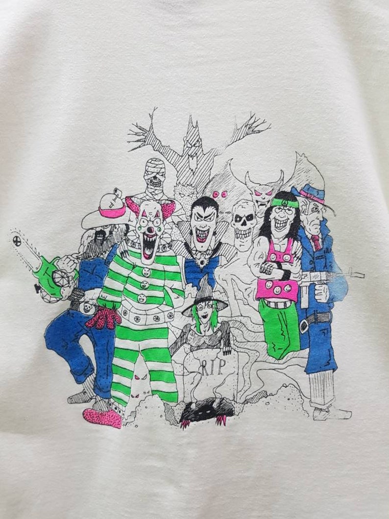Puede incluir: Una camiseta blanca con una ilustraci&oacute;n en blanco y negro de un grupo de personajes espeluznantes. Los personajes incluyen un payaso, un vampiro, una bruja, un zombi y un monstruo. La ilustraci&oacute;n est&aacute; rodeada de un fondo blanco y negro con un &aacute;rbol y una l&aacute;pida que dice "RIP".