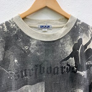 May include: Gray t-shirt with a black and white all-over print of a skateboarder. The shirt has a white crew neck and a "CALAJA" label on the front.