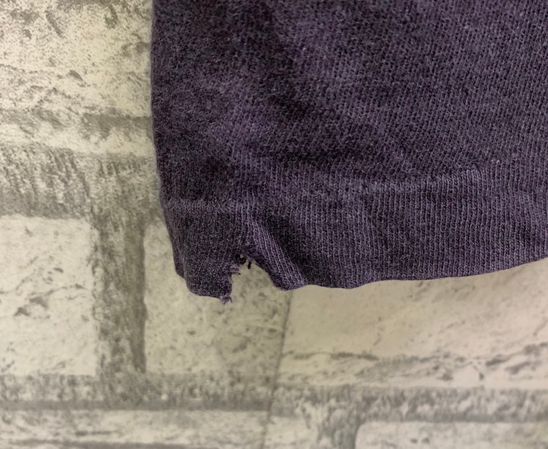 May include: Close-up of the bottom edge of a dark purple t-shirt. The fabric is soft and has a slight texture. The edge of the shirt is frayed.