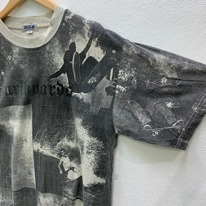 May include: Gray t-shirt with a faded print of skateboarders and waves. The shirt has a crew neck and short sleeves. The text "CALI-JA" is visible on the tag.