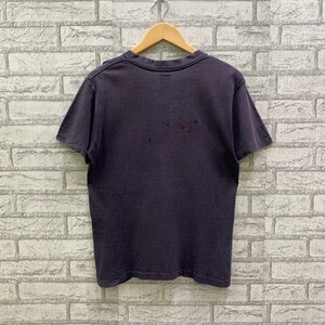May include: A dark gray t-shirt with short sleeves and a crew neck. The shirt is hanging on a wooden hanger against a white brick wall.