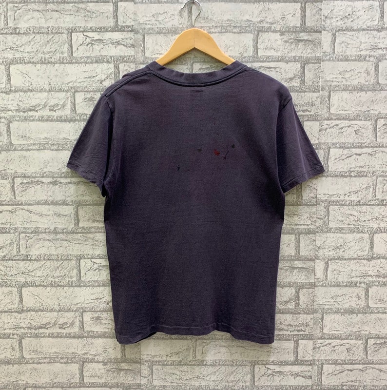 May include: A dark gray t-shirt with short sleeves and a crew neck. The shirt is hanging on a wooden hanger against a white brick wall.