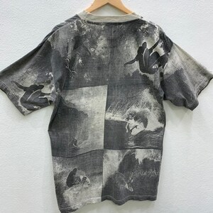 May include: Gray short-sleeved t-shirt with a faded print of surfers riding waves. The print is in a grid pattern with four squares.