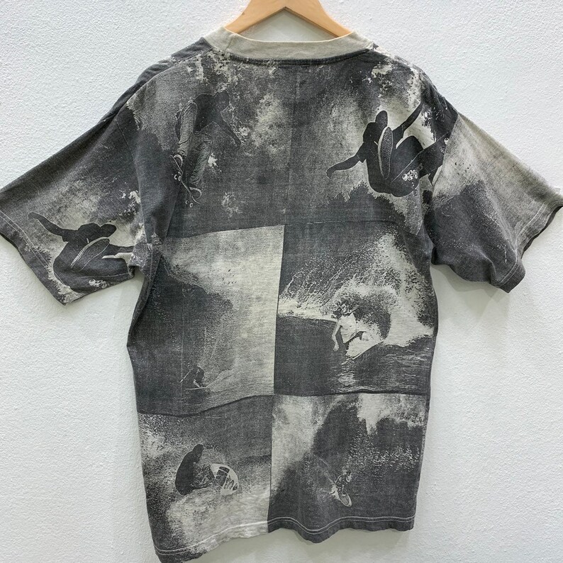 May include: Gray short-sleeved t-shirt with a faded print of surfers riding waves. The print is in a grid pattern with four squares.