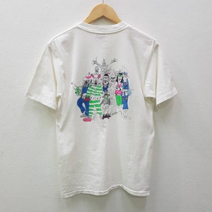 Puede incluir: Camiseta blanca con un estampado gr&aacute;fico de un grupo de personajes de dibujos animados en blanco y negro con detalles verdes y azules. Los personajes llevan varios disfraces y sostienen objetos.