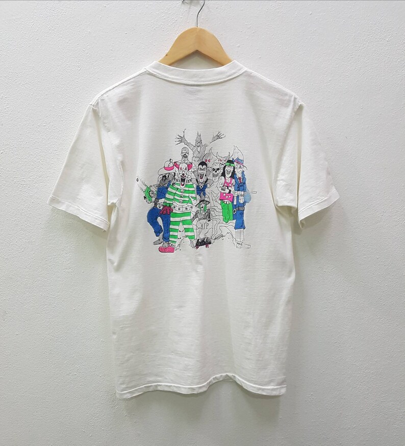 Puede incluir: Camiseta blanca con un estampado gr&aacute;fico de un grupo de personajes de dibujos animados en blanco y negro con detalles verdes y azules. Los personajes llevan varios disfraces y sostienen objetos.