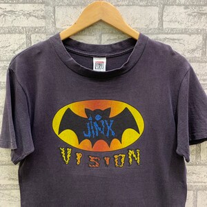 May include: A dark purple t-shirt with a yellow and orange bat graphic. The bat has a blue "Jinx" written on it. The text "Vision" is written in a stylized font below the bat. The t-shirt has a white tag that says "Vision Street Wear" on it.