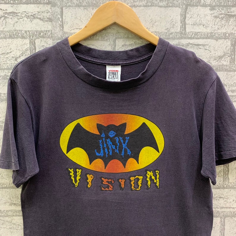 May include: A dark purple t-shirt with a yellow and orange bat graphic. The bat has a blue "Jinx" written on it. The text "Vision" is written in a stylized font below the bat. The t-shirt has a white tag that says "Vision Street Wear" on it.