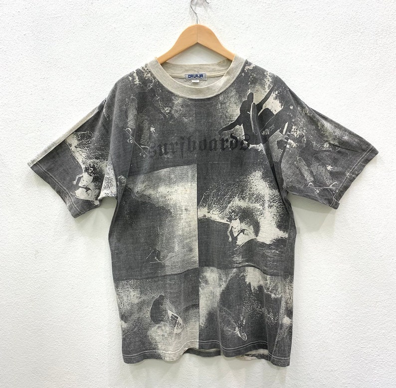 May include: A gray t-shirt with a faded all-over print of surfers and waves. The text "Surfboards" is printed in black on the front of the shirt.