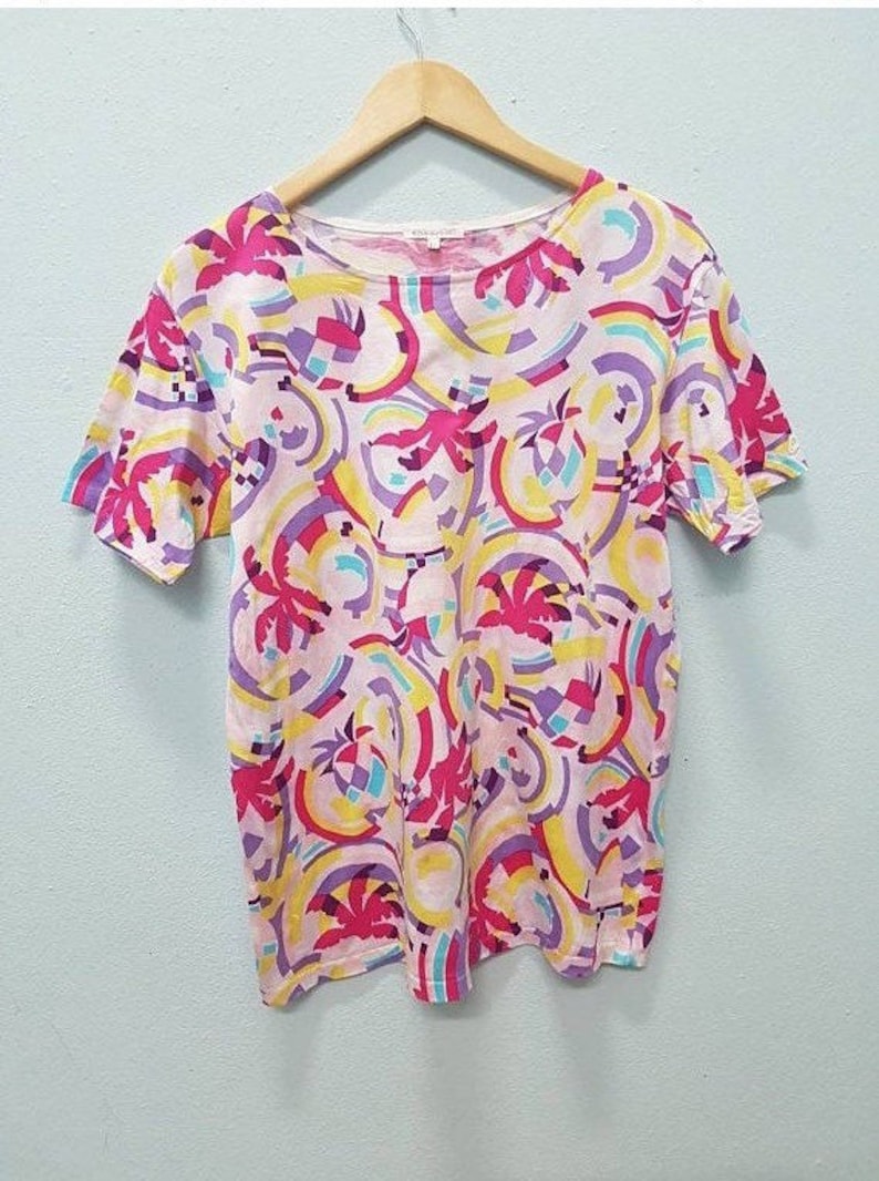 May include: A pink short-sleeved t-shirt with a colorful abstract print featuring palm trees, circles, and geometric shapes in shades of pink, yellow, blue, and purple.