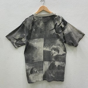 May include: A gray short-sleeved t-shirt with a black and white print of surfers riding waves. The print is repeated across the entire shirt.