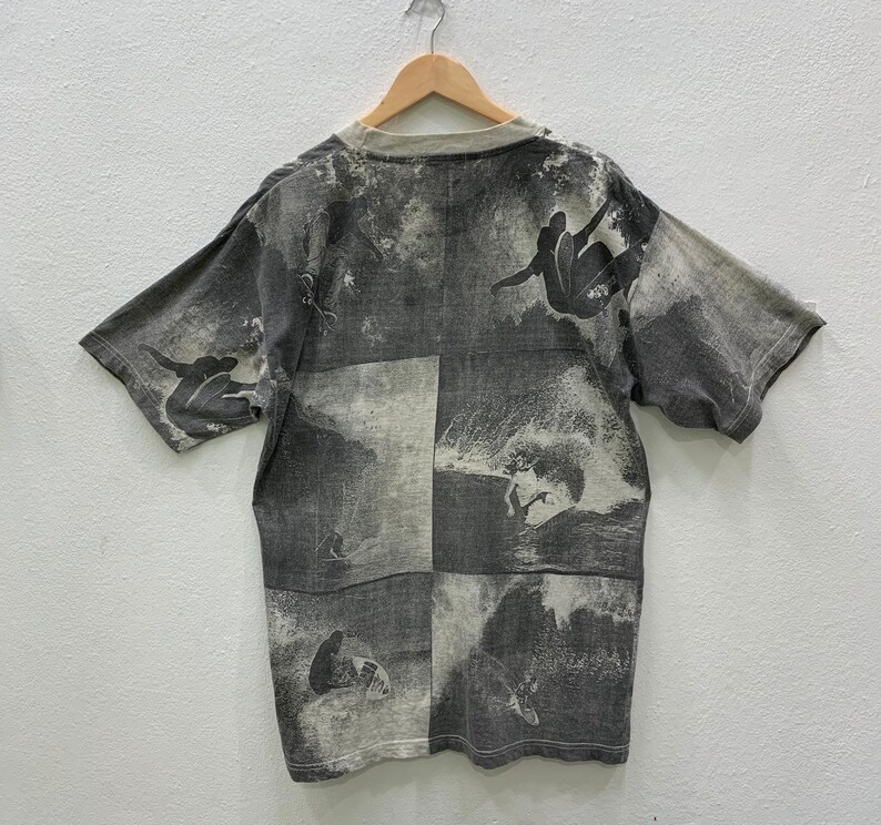 May include: A gray short-sleeved t-shirt with a black and white print of surfers riding waves. The print is repeated across the entire shirt.