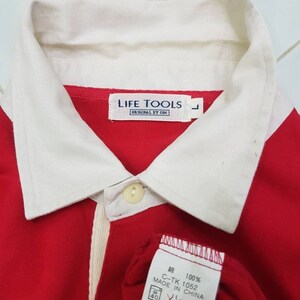 May include: Red and white long-sleeve shirt with a white collar and a white stripe down the front. The shirt has a button closure and a label that reads "LIFE TOOLS ORIGINAL BY COK" and "100% 綿 C-TK 1052 MADE IN CHINA".