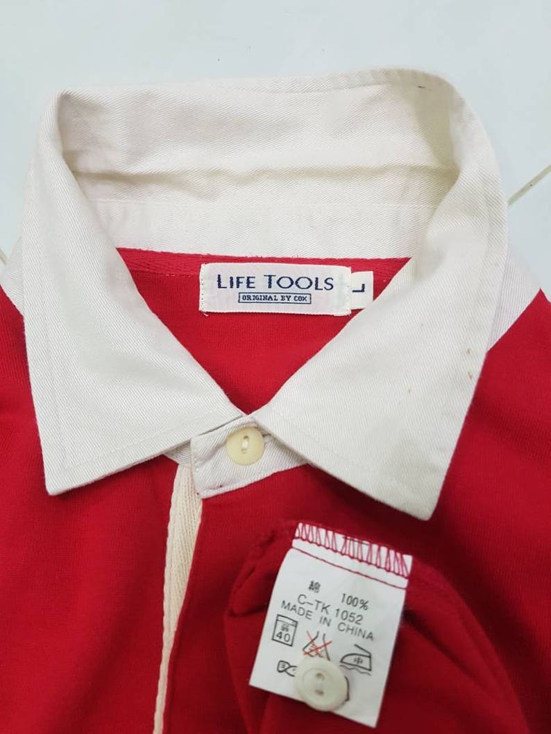 May include: Red and white long-sleeve shirt with a white collar and a white stripe down the front. The shirt has a button closure and a label that reads "LIFE TOOLS ORIGINAL BY COK" and "100% 綿 C-TK 1052 MADE IN CHINA".