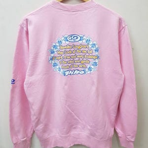 May include: A light pink sweatshirt with a crew neck and long sleeves. The back features a floral design with text that reads "Hawaiian Longboard Wear Look at the sea full of spirit, true and easy footsteps of the wind and gentle touch of the earth. Pipe."