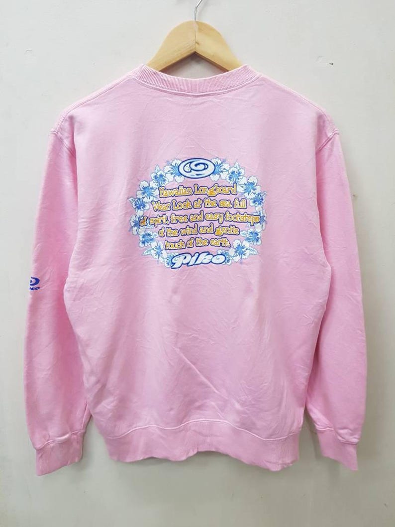 May include: A light pink sweatshirt with a crew neck and long sleeves. The back features a floral design with text that reads "Hawaiian Longboard Wear Look at the sea full of spirit, true and easy footsteps of the wind and gentle touch of the earth. Pipe."