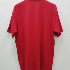 May include: A red short-sleeved polo shirt with a beige collar and a button closure. The shirt has a plain design and is hanging on a hanger.