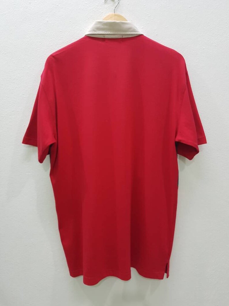 May include: A red short-sleeved polo shirt with a beige collar and a button closure. The shirt has a plain design and is hanging on a hanger.