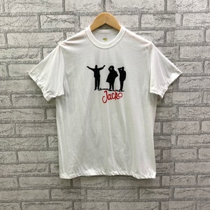 Peut inclure: T-shirt blanc avec une silhouette noire de trois personnes et le mot "Jacko" en lettres rouges.