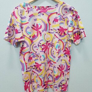 May include: A pink, yellow, purple, and blue short-sleeved shirt with a geometric pattern. The shirt has a scoop neck and a relaxed fit.