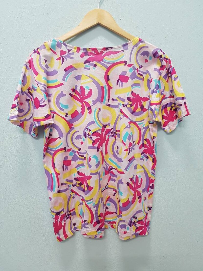 May include: A pink, yellow, purple, and blue short-sleeved shirt with a geometric pattern. The shirt has a scoop neck and a relaxed fit.