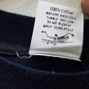 May include: White care label with black text that reads "100% Cotton Machine Wash Cold Tumble Dry Low Do Not Bleach Wash Separately". A black and white illustration of a skeleton surfing is below the text.