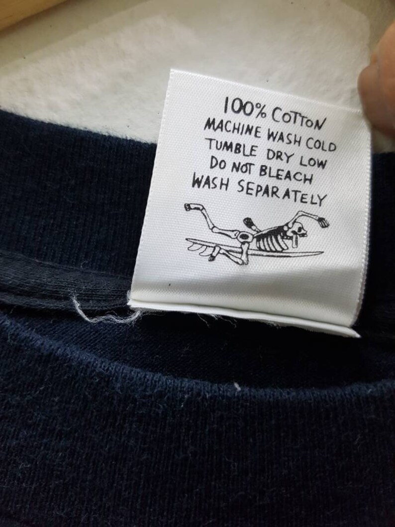 May include: White care label with black text that reads "100% Cotton Machine Wash Cold Tumble Dry Low Do Not Bleach Wash Separately". A black and white illustration of a skeleton surfing is below the text.