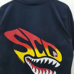 May include: Black t-shirt with a red, yellow, and white graphic of a shark with teeth and an eye. The text "S.C.O.P." is printed on the graphic. The copyright symbol and "Sally Ann Designs" are printed below the graphic.