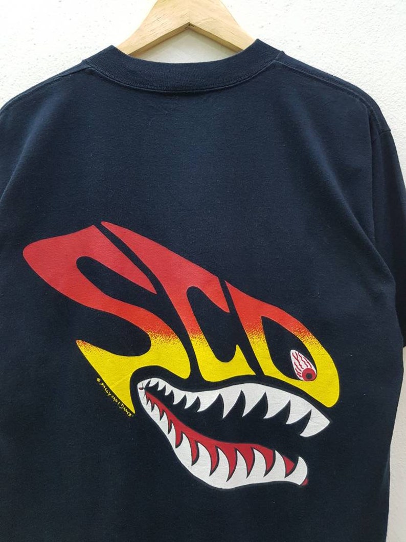 May include: Black t-shirt with a red, yellow, and white graphic of a shark with teeth and an eye. The text "S.C.O.P." is printed on the graphic. The copyright symbol and "Sally Ann Designs" are printed below the graphic.