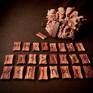 Peut inclure: Un ensemble de tuiles rectangulaires en bois gravées de symboles runiques, disposées en rangées. Un pendentif rectangulaire en bois avec un motif quadrillé est suspendu à une pochette en crochet beige avec un cordon en cuir marron.