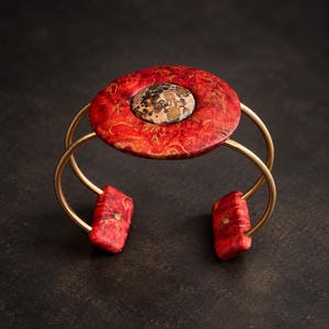 Könnte beinhalten: Ein Armreif mit einem kreisförmigen Mittelstück aus rot-orange marmoriertem Holz und einer zentralen Einlage aus gesprenkeltem braunem Stein. Der Armreif besteht aus zwei dünnen goldfarbenen Metallbändern, die mit zwei rechteckigen roten Holzstücken verbunden sind.