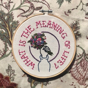 May include: Embroidered hoop art featuring the question "WHAT IS THE MEANING OF LIFE?" in pink thread. A hand holding a bouquet of flowers is embroidered in the centre. The flowers are in shades of pink, blue, and purple.
