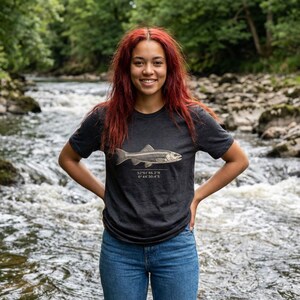 Puede incluir: Camiseta gris oscuro con una ilustración detallada de un esqueleto de pez y coordenadas debajo. La camiseta se combina con unos vaqueros azules.