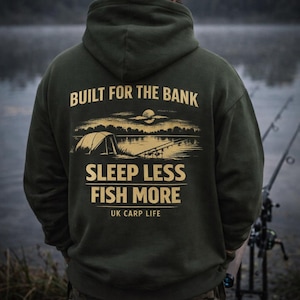 May include: Dark green hoodie with the text "BUILT FOR THE BANK" and "SLEEP LESS FISH MORE" in gold lettering. The back of the hoodie features a graphic of a lakeside scene with a tent and fishing rods. The text "UK CARP LIFE" is also included.