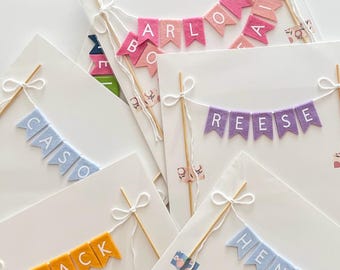 May include: Multiple felt cake toppers featuring individual pennant flags strung on white twine between two wooden skewers. The flags display various names and phrases including Navy Bean, Happy Birthday, Arlons Air, Reese, Jack, and Henry in colors.