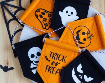 May include: A collection of five small felt pennants in black and orange, featuring Halloween designs including a ghost, a skull, a pumpkin, a jack-o'-lantern face, and the text "TRICK or TREAT" with a bat icon, all suspended by black and white twine.