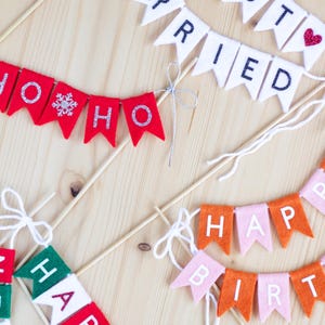 May include: Multiple felt pennant banners on wooden sticks. One white banner reads JUST MARRIED with a red heart. A red banner reads HO HO HO with a silver snowflake. A pink and orange banner reads HAPPY BIRTHDAY. A red, white, and green banner reads HAPPY.