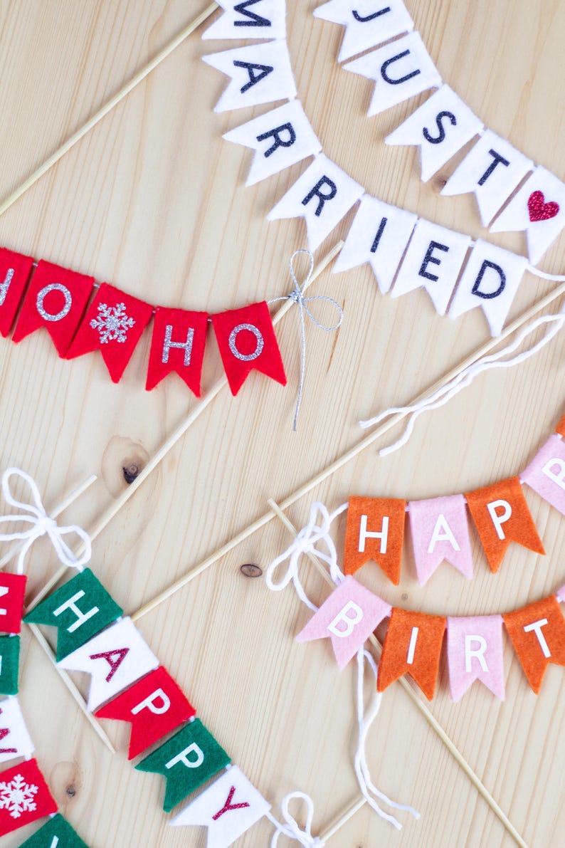 May include: Multiple felt pennant banners on wooden sticks. One white banner reads JUST MARRIED with a red heart. A red banner reads HO HO HO with a silver snowflake. A pink and orange banner reads HAPPY BIRTHDAY. A red, white, and green banner reads HAPPY.