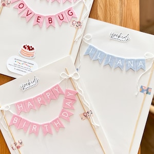 May include: Three handmade felt bunting cake toppers on white backing cards. One reads WELCOME LOVE BUG in pink, one reads ILIANA in light blue, and one reads HAPPY BIRTHDAY in pink. Each features wooden skewers, white string bows, and branding.