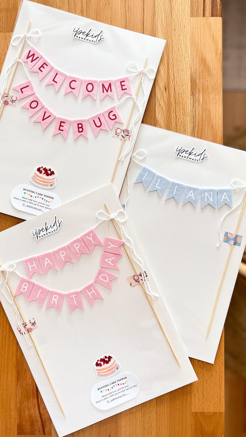 May include: Three handmade felt bunting cake toppers on white backing cards. One reads WELCOME LOVE BUG in pink, one reads ILIANA in light blue, and one reads HAPPY BIRTHDAY in pink. Each features wooden skewers, white string bows, and branding.